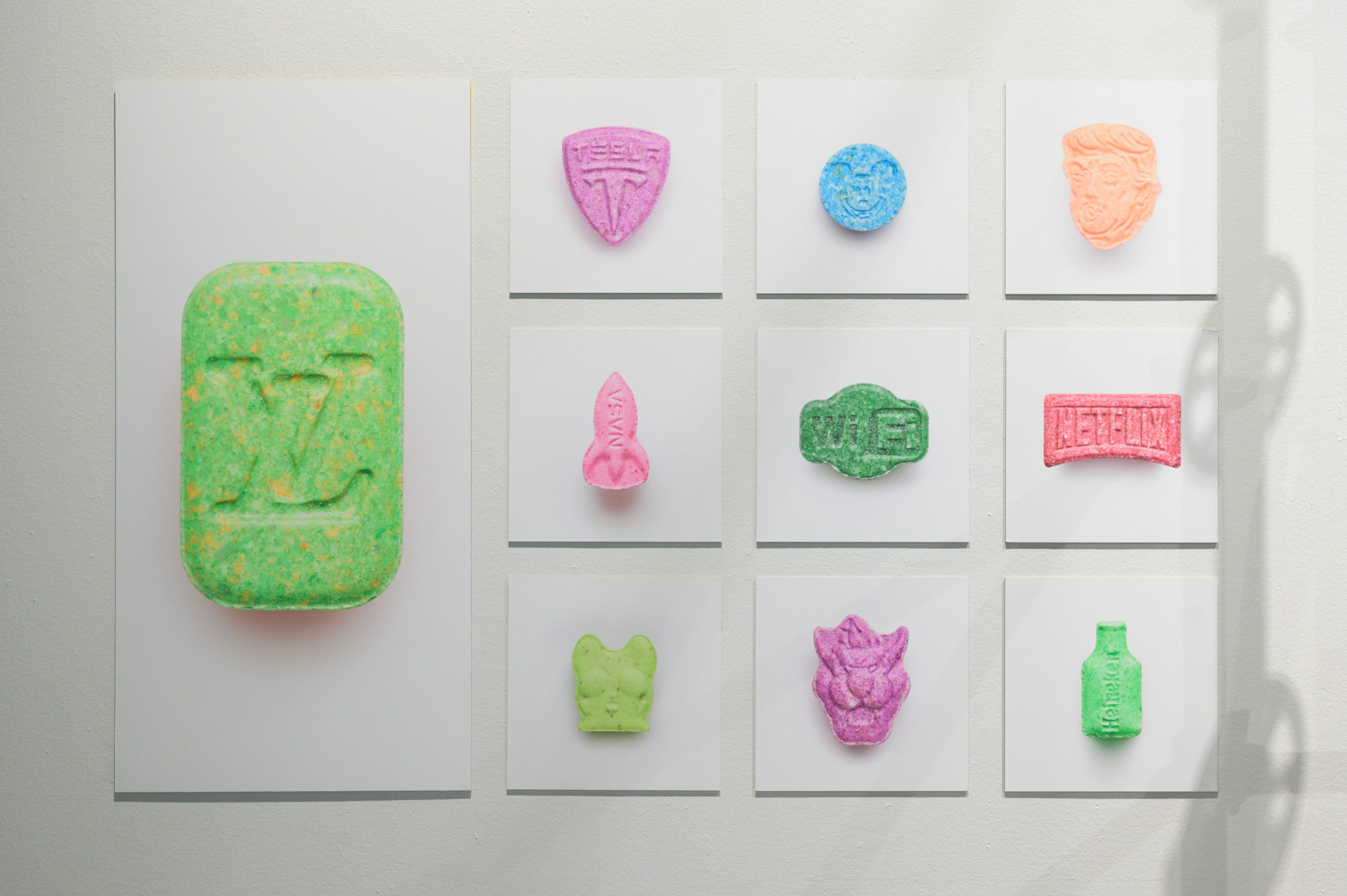 Een foto van 10 foto's aan een muur waarop steeds één xtc-pil te zien is op een witte achtergrond.