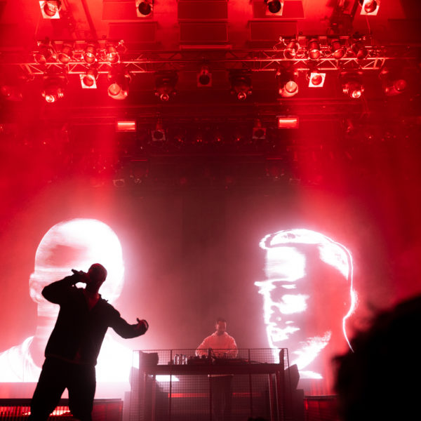 Optreden van The Opposites. De frontman staat op het podium met een microfoon, achter hem op het scherm zijn twee grafische gezichten te zien.