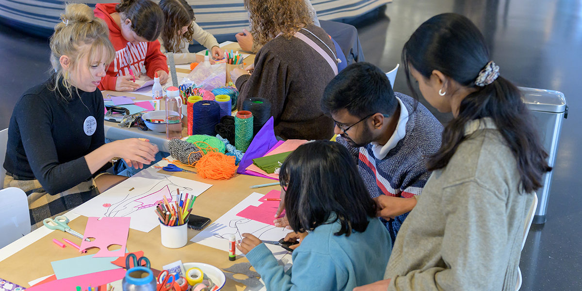 Een gezin met moeder, vader en kind zitten aan tafel te knutselen. De museumdocent zit er tegenover en knutselt mee.
