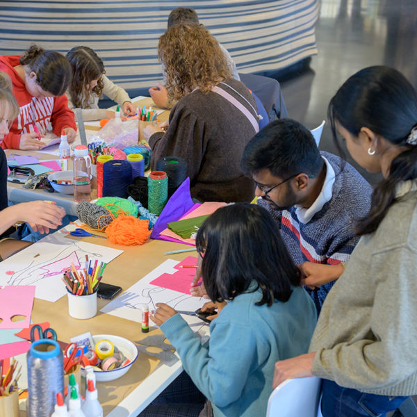 Een gezin met moeder, vader en kind zitten aan tafel te knutselen. De museumdocent zit er tegenover en knutselt mee.