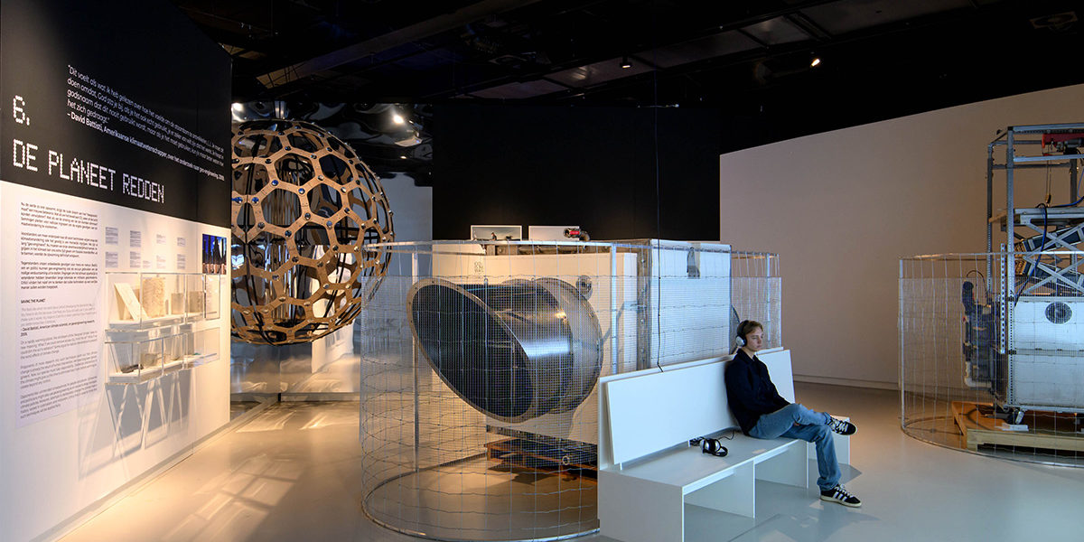 In een tentoonstellingszaal zit een man op een bankje. Achter hem staat een grote, zilverkleurige co2-afvangmachine.