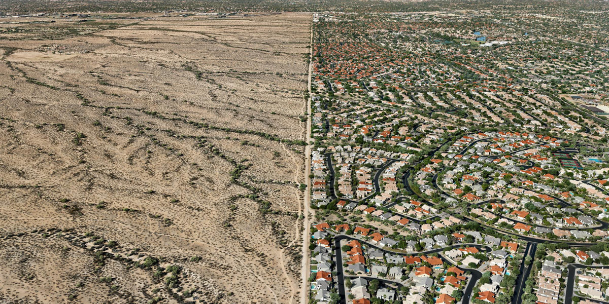 Een landschap van boven gefotografeerd. Links de droge vlakte van een reservaat. In het midden loop een rechte lijn, met rechts daarvan een drukbevolkte stad met laagbouw en bomen.