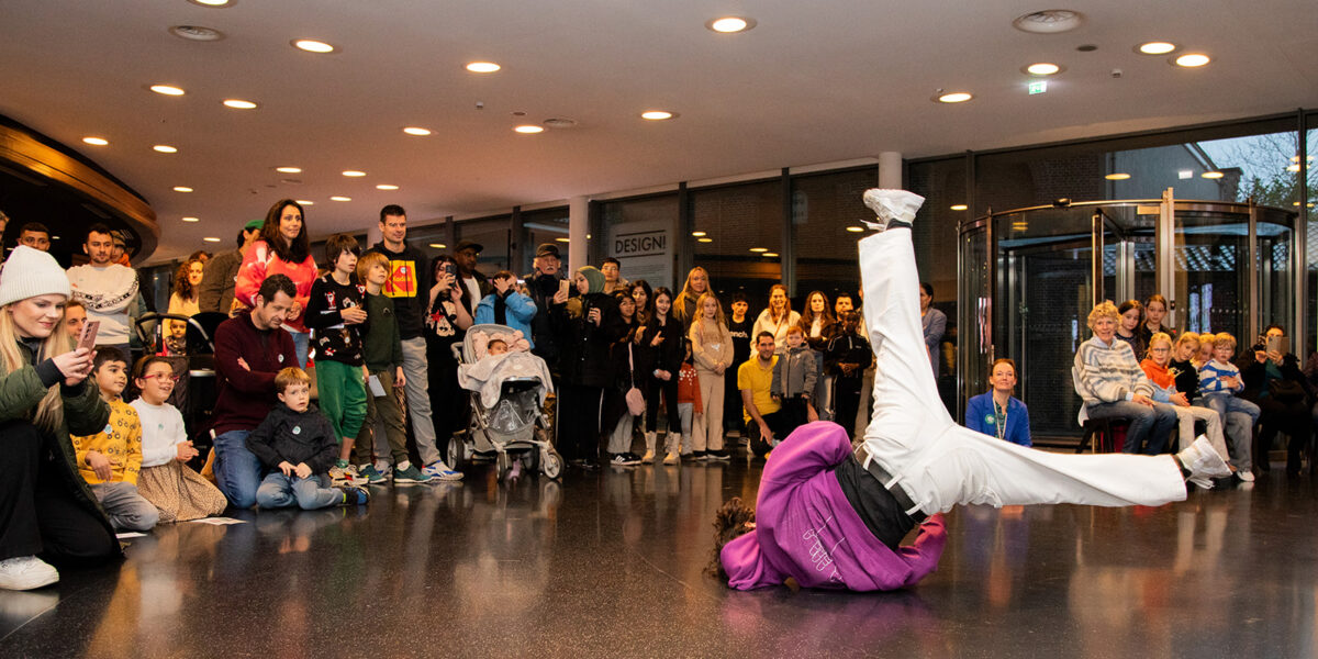 Een foto van een breakdancende jongen die op zijn kop draait, met daaromheen een publiek met veel verschillende mensen.